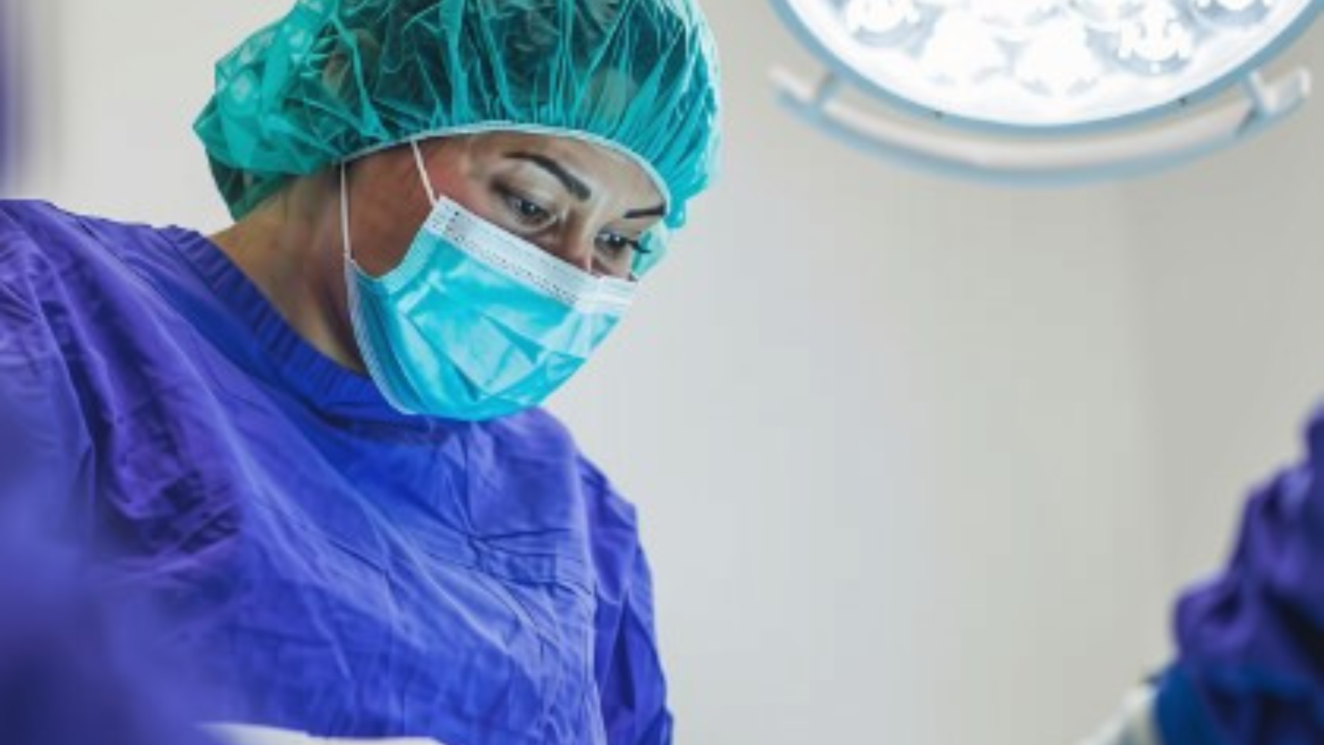Close-up of a healthcare professional in sterile surgical attire performing a procedure in an operating room, illuminated by overhead surgical lighting.