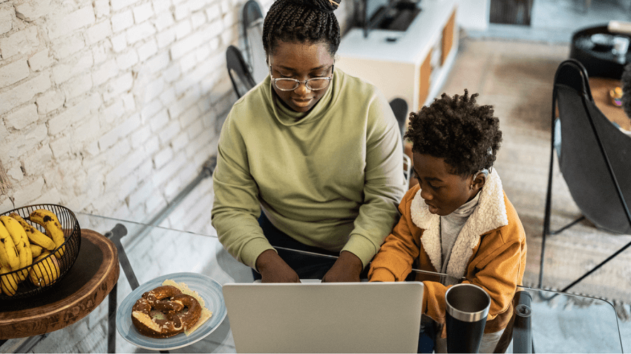 An adult and child sitting together at a table using a laptop. The child is wearing a hearing aid.