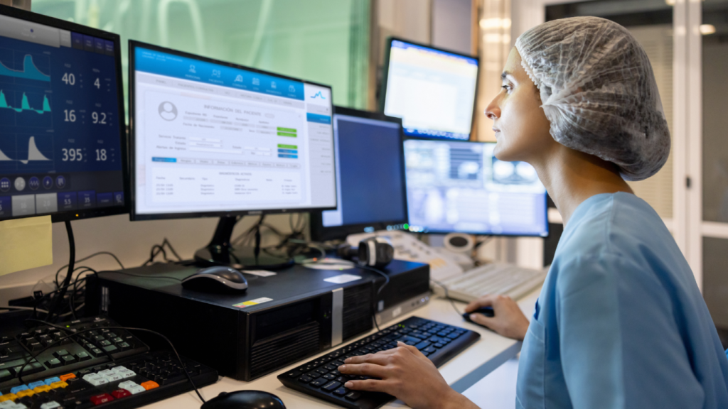 Healthcare professional in a clinical setting working at a workstation with multiple monitors displaying patient information, medical data, and diagnostic dashboards. T