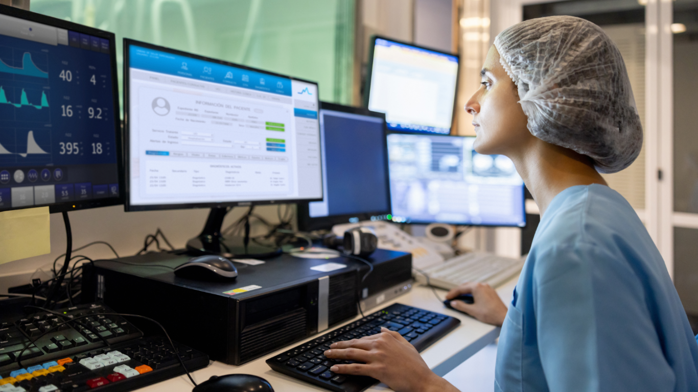 Healthcare professional in a clinical setting working at a workstation with multiple monitors displaying patient information, medical data, and diagnostic dashboards. T