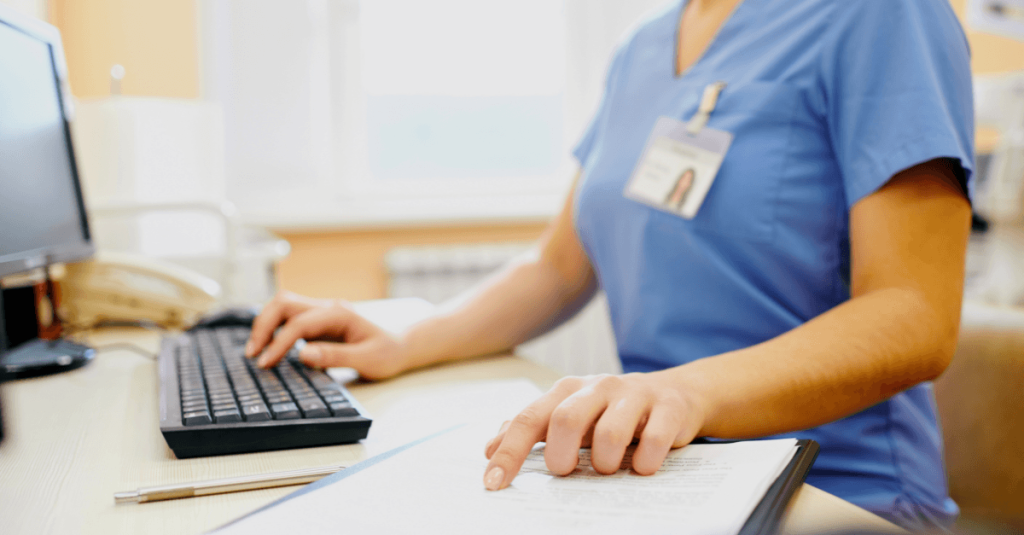 Healthcare professional in scrubs reviews patient documents while using a computer keyboard at a medical office desk.