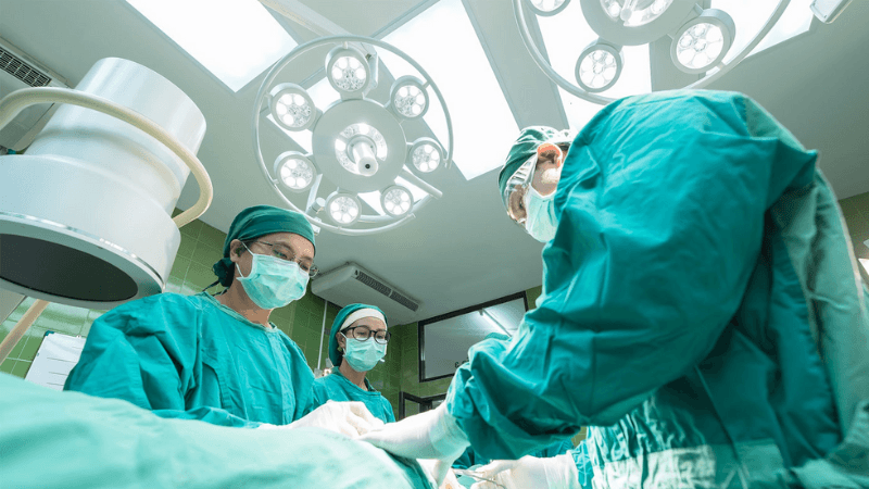 Surgical team in an operating room performs a procedure under bright overhead lights while wearing protective gear.