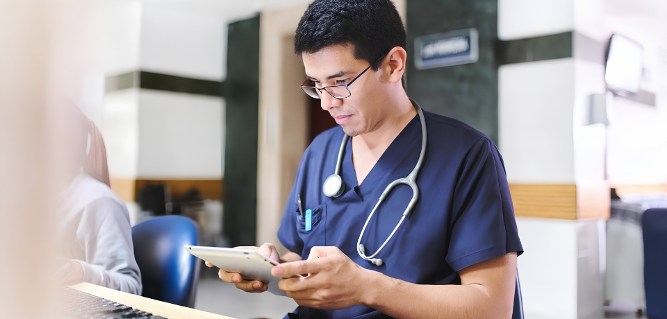 Medical staff member in a clinical setting checks patient information on a tablet, with a stethoscope visible.
