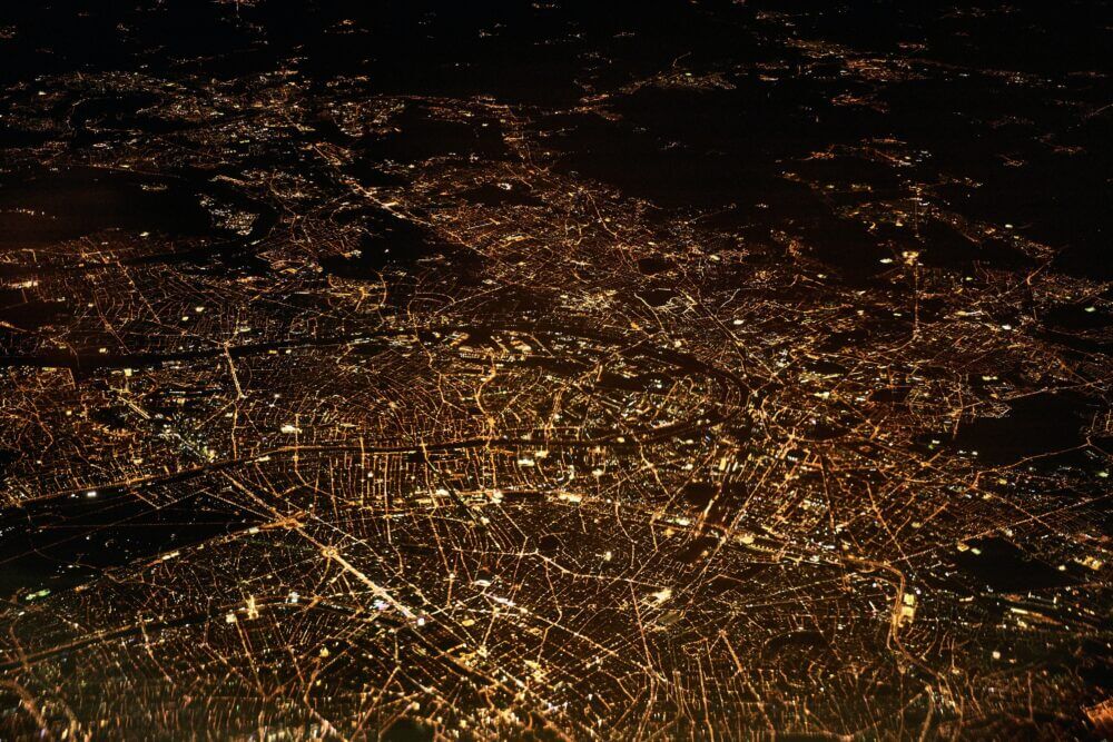 Aerial nighttime view of a brightly illuminated cityscape, showing connected roads, neighborhoods, and urban networks.