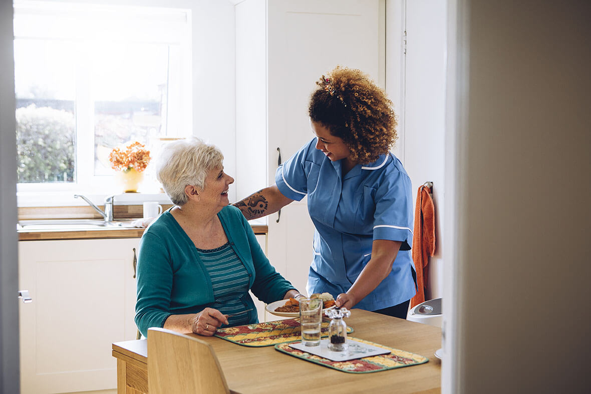 Home care worker assisting an older adult during mealtime in a domestic kitchen.