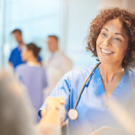 Healthcare professional in scrubs greets a visitor while speaking in a bright clinical setting with staff nearby.