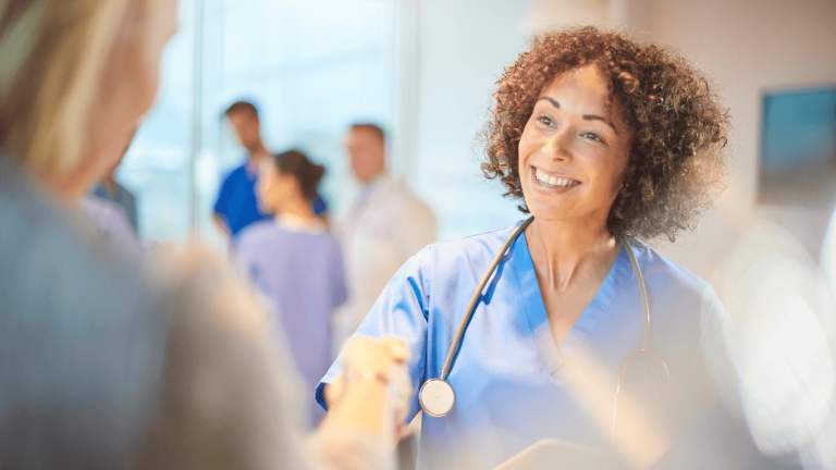 Healthcare professional in scrubs greets a visitor while speaking in a bright clinical setting with staff nearby.