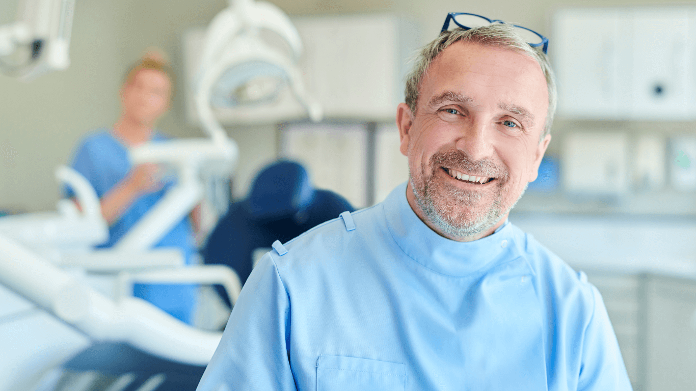 Person in a blue clinical uniform in a modern dental treatment room, with dental equipment visible in the background.