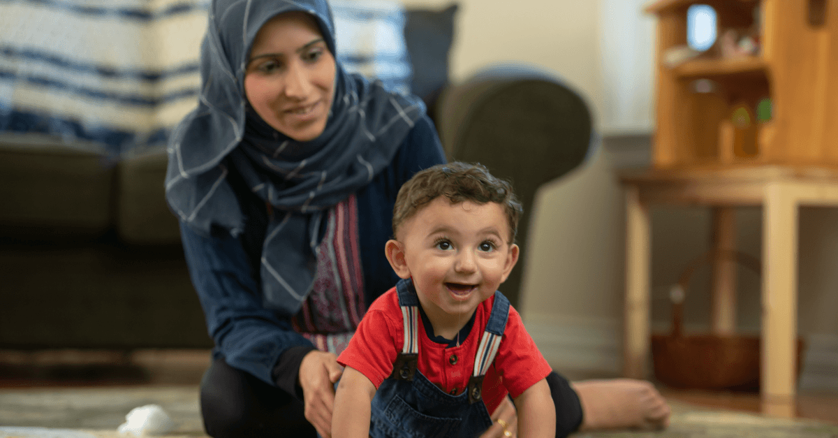 Young child in red overalls crawls across a living room floor while a caregiver sits nearby, offering support in a home setting.