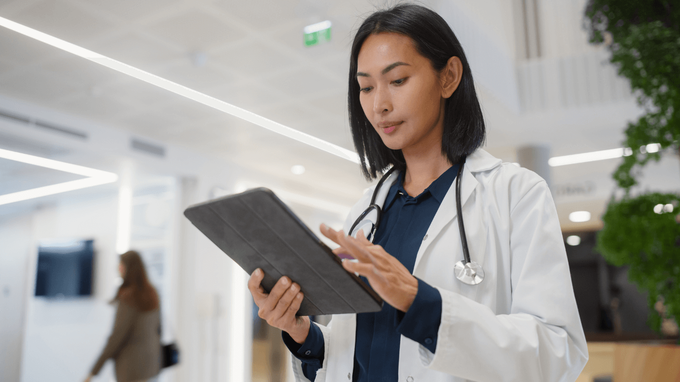 Healthcare professional in a clinical setting reviews information on a tablet while wearing a lab coat and stethoscope.