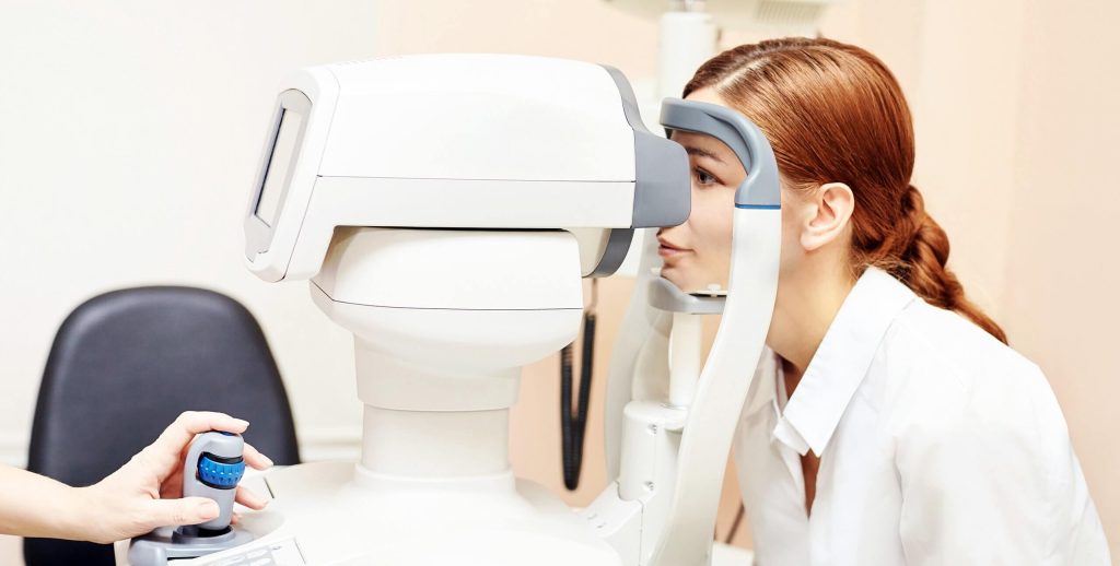 Person undergoing an eye examination using an automated retinal imaging device, while a clinician operates the testing equipment.