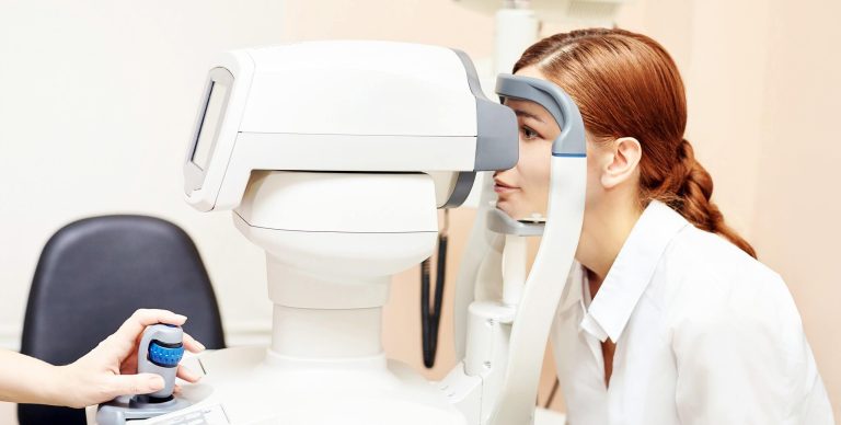Person undergoing an eye examination using an automated retinal imaging device, while a clinician operates the testing equipment.