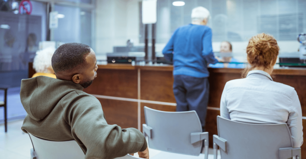 Patients sit in a healthcare waiting area while another person speaks with staff at the reception desk in a modern clinic setting.