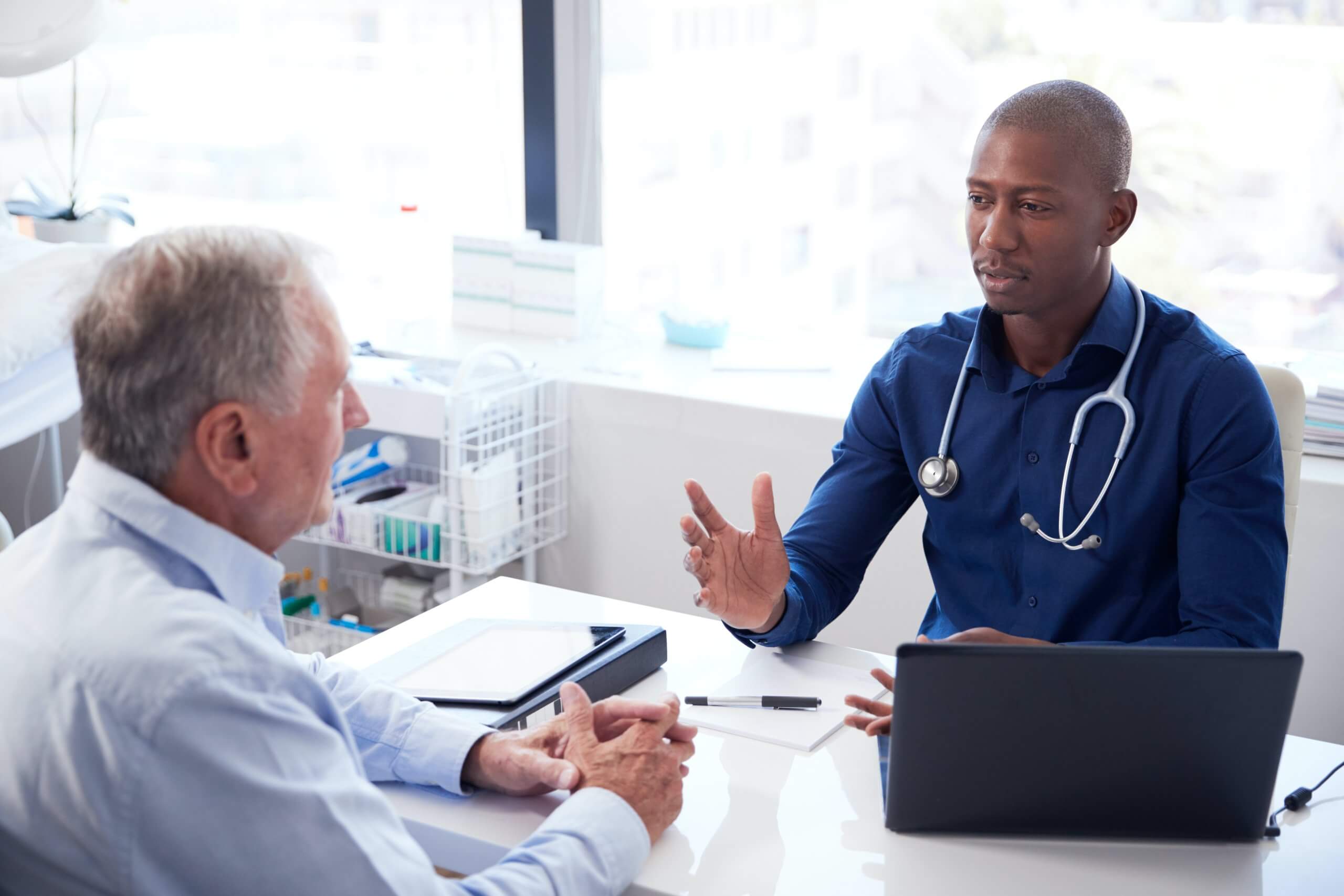 Healthcare professional consulting with an older patient across a desk in a bright clinic, with a laptop and notes visible.