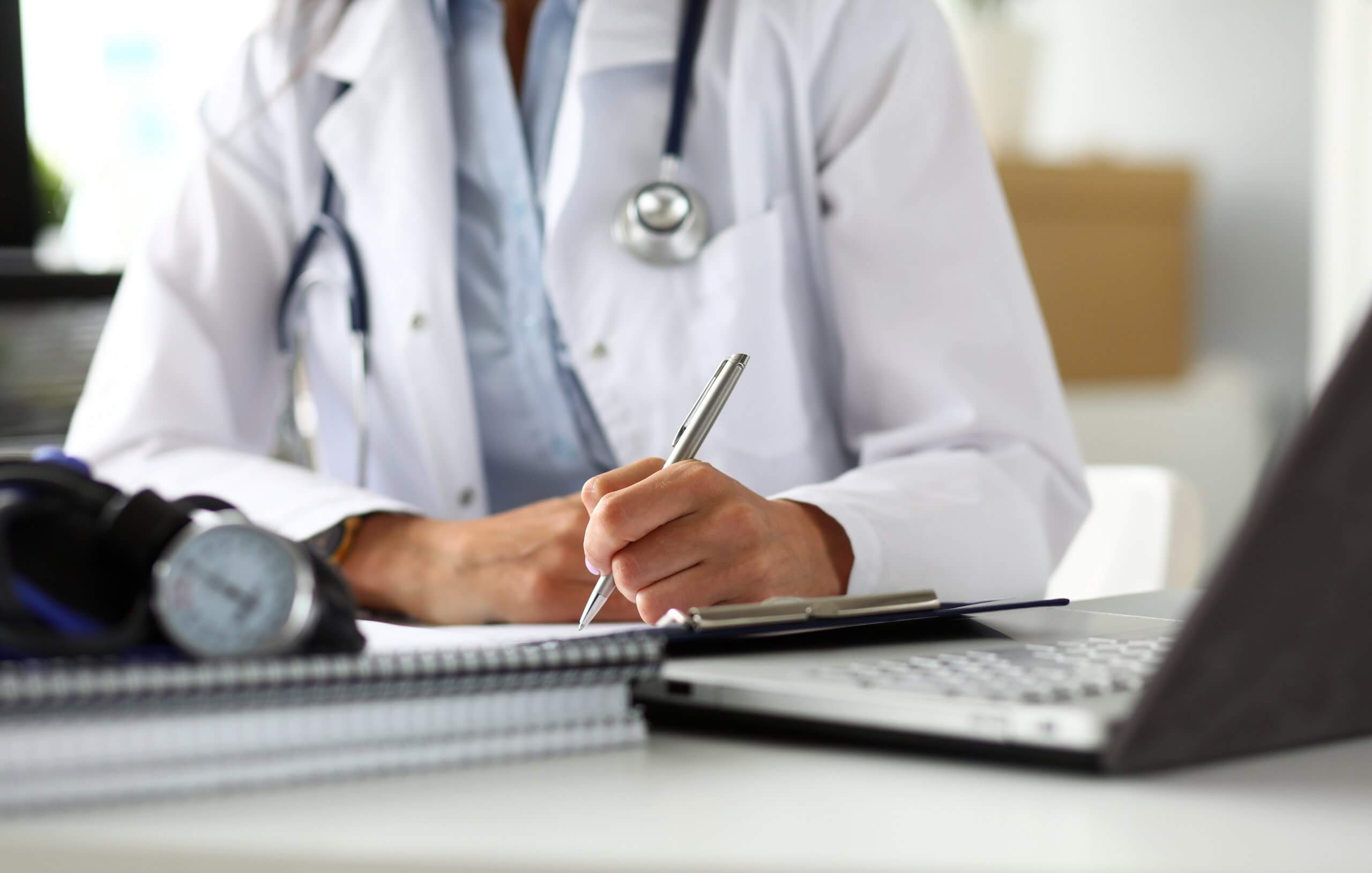 Close-up of a healthcare professional documenting information on a clipboard in a clinical office setting, with a laptop, stethoscope and blood pressure monitor visible in the foreground.