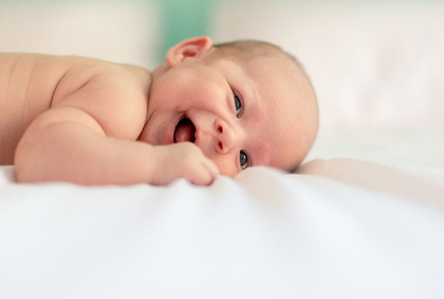 Newborn baby lying on a soft white blanket with one hand near the face, looking toward the camera.