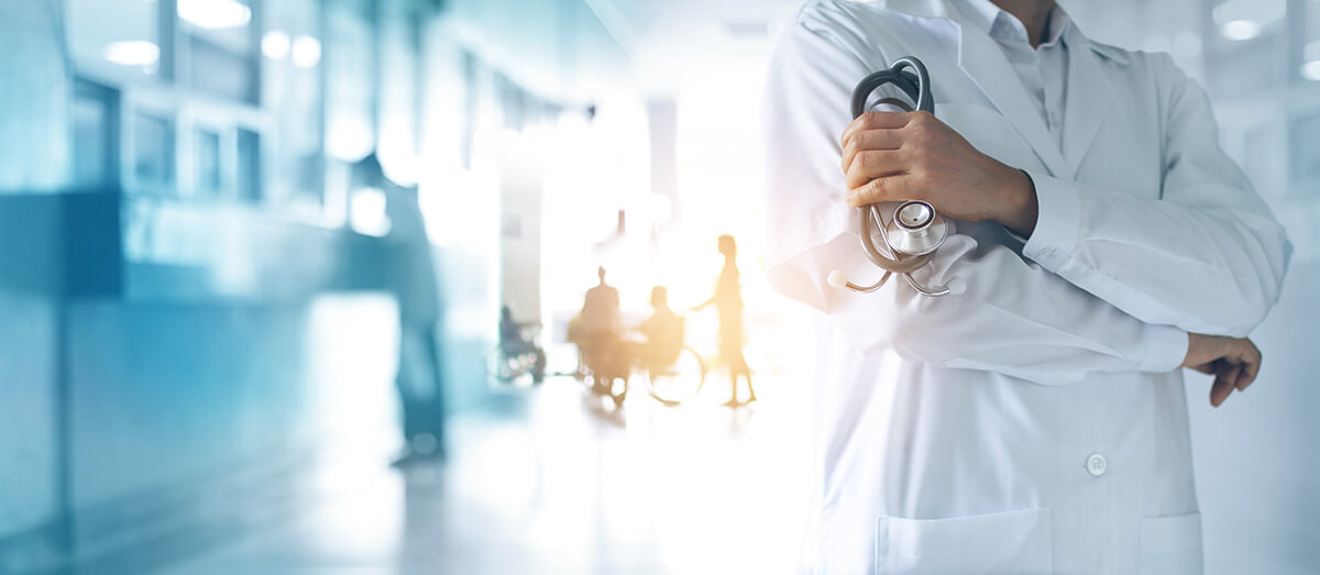 Healthcare professional holding a stethoscope in a modern hospital corridor, with healthcare activity visible in the background.