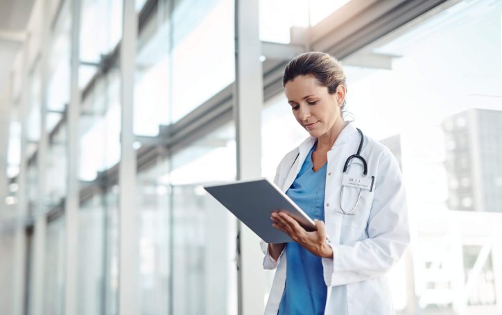 Healthcare professional in a white coat uses a tablet to review patient information in a bright hospital corridor with large windows.