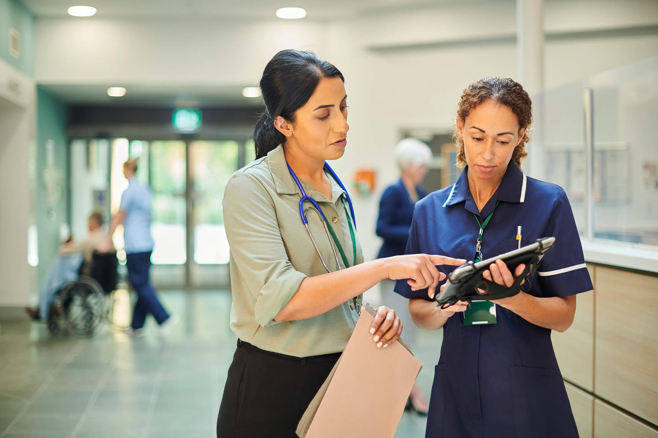 Two healthcare professionals standing in a hospital corridor, discussing information displayed on a tablet device