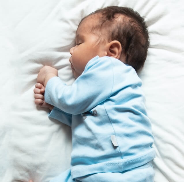 Sleeping newborn in a light blue outfit lies on a white blanket with hands clasped.